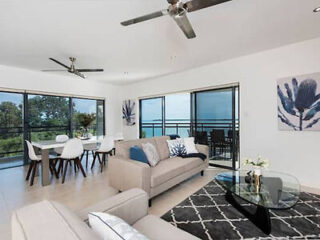 Modern living room with a dining area, featuring large windows overlooking the ocean. The room is furnished with a grey sofa, glass coffee table, and a dining table with white chairs, and ceiling fans.