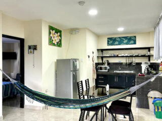 A modern kitchen and dining area with a steel refrigerator, black cabinets, and a hanging blue hammock. The space is decorated with framed art and includes a table with four chairs.