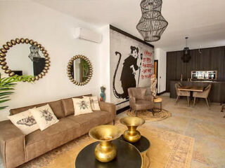 A modern living room with a brown sofa, decorative mirrors, gold coffee tables, and wall art of a large rat. There is a dining area with a wooden table and chairs in the background.