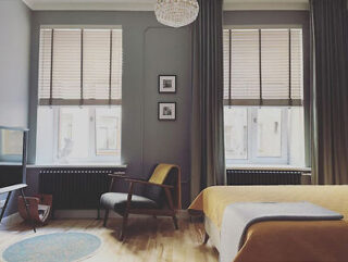 A modern bedroom with a single bed, wooden chair, two large windows with blinds, framed pictures on the wall, a chandelier, and a light wooden floor with a small rug.