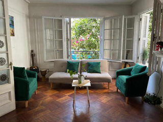 Bright living room with a light sofa, green armchairs, and large windows overlooking lush greenery.