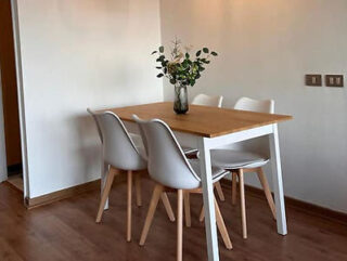 Small dining area with a wooden table, four white chairs, and a vase of flowers, set against a simple wall.