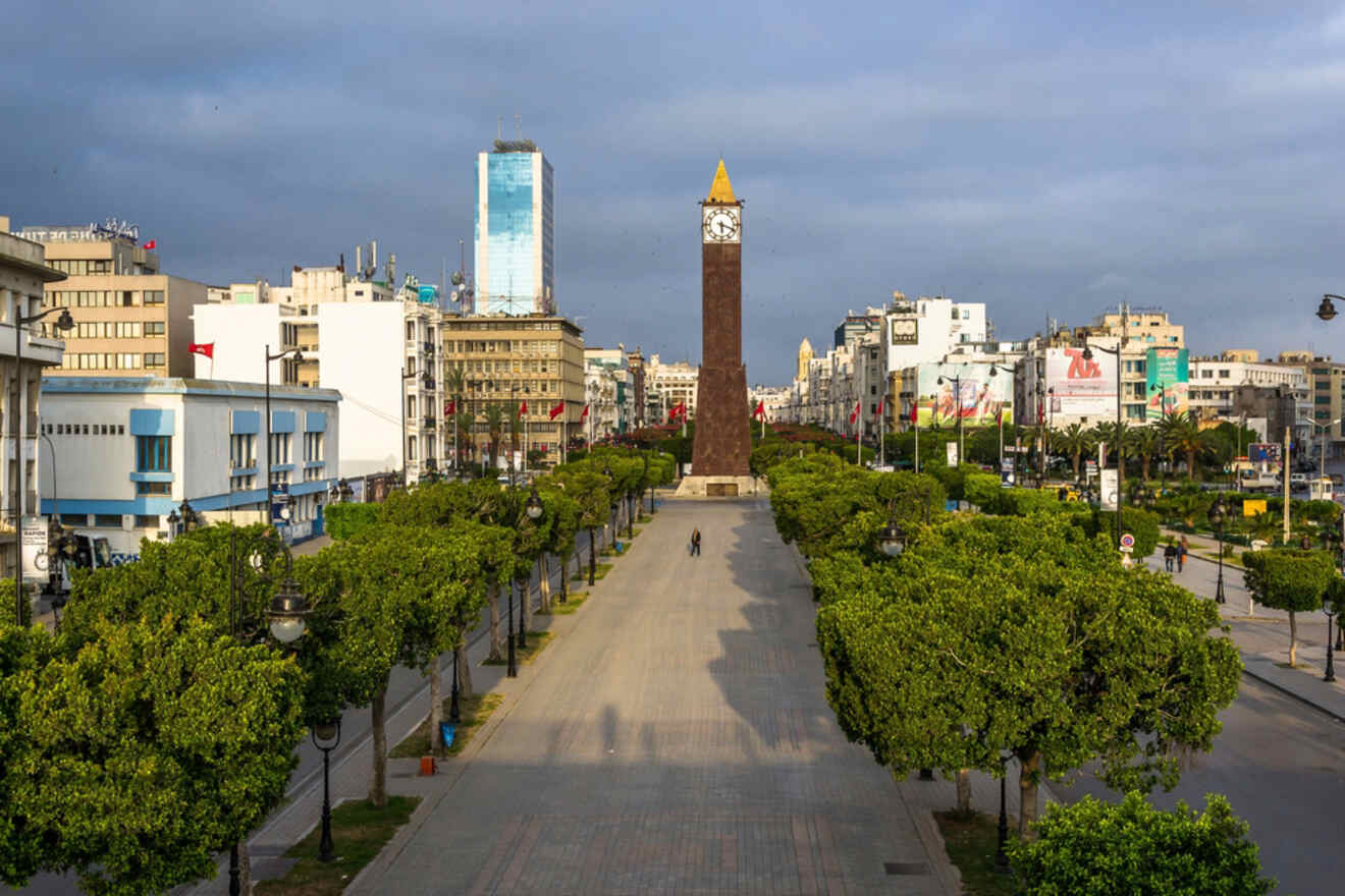 A wide boulevard in downtown Tunis, with a tall clock tower at the center and tree-lined sidewalks leading towards modern office buildings.