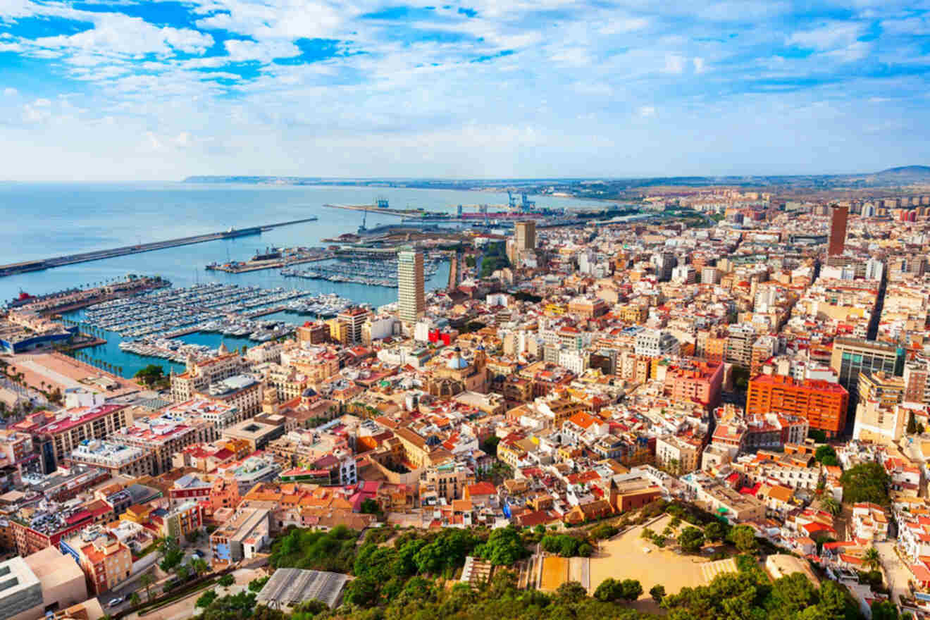 Aerial view of a coastal city with a marina, numerous buildings, and greenery under a partly cloudy sky.