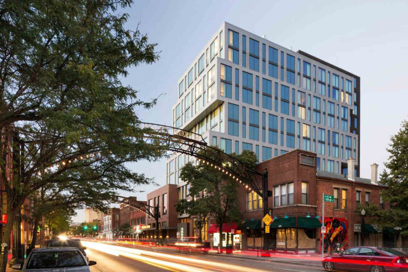 Street view of a mixed-use building with modern glass and concrete design, adjacent to older brick buildings. Cars leave light trails along the road, and a decorative arch stretches over the street.