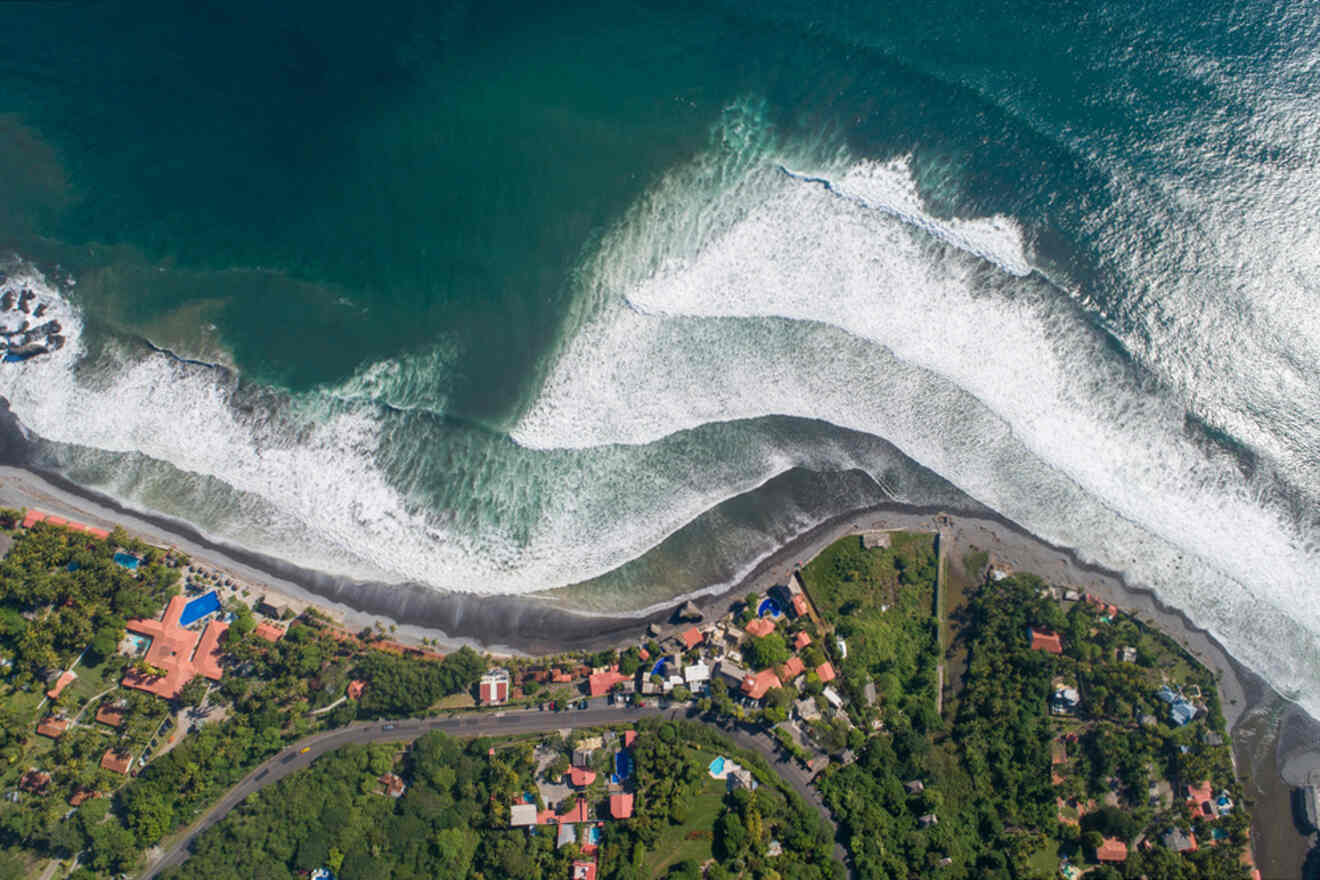 Aerial view of a coastal area with waves crashing on a sandy beach, adjacent to a road lined with houses and greenery.