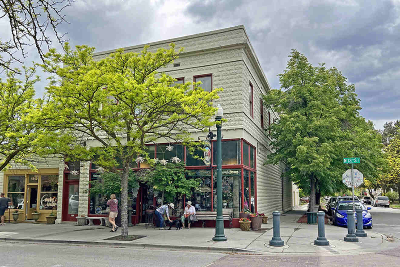 A street corner with a two-story building, trees, people sitting on a bench, and a small car parked nearby under a cloudy sky.