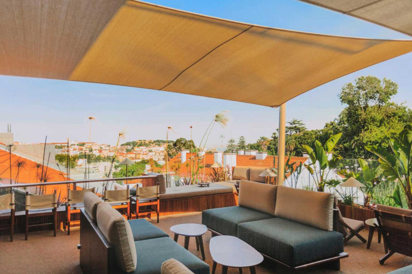 Outdoor lounge area with modern sofas and small tables under a large canopy. The space overlooks a town with red-roofed buildings and greenery in the distance.