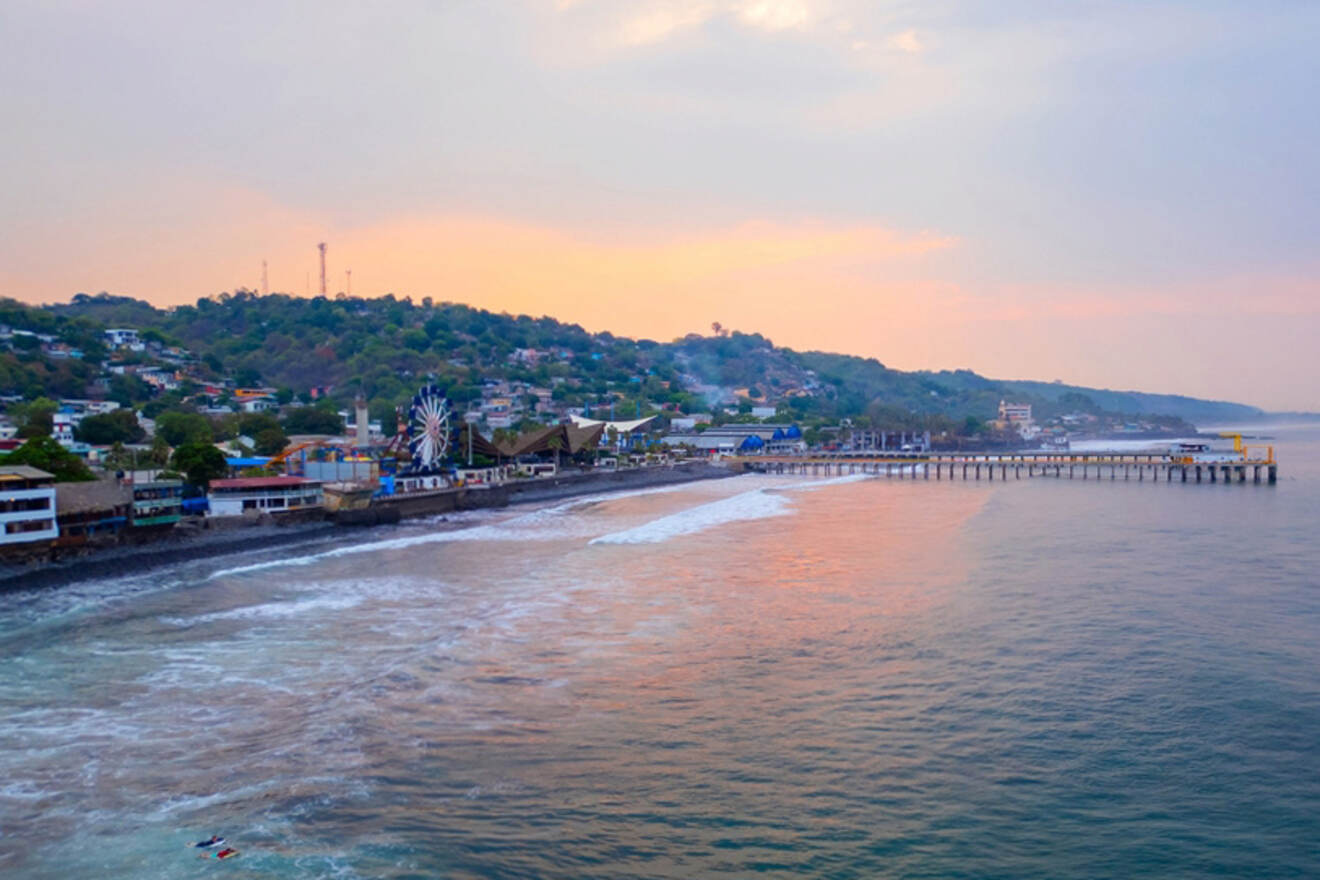 A seaside town with a pier extending into the ocean, surrounded by hills and buildings, under a pastel-colored sky at sunset.