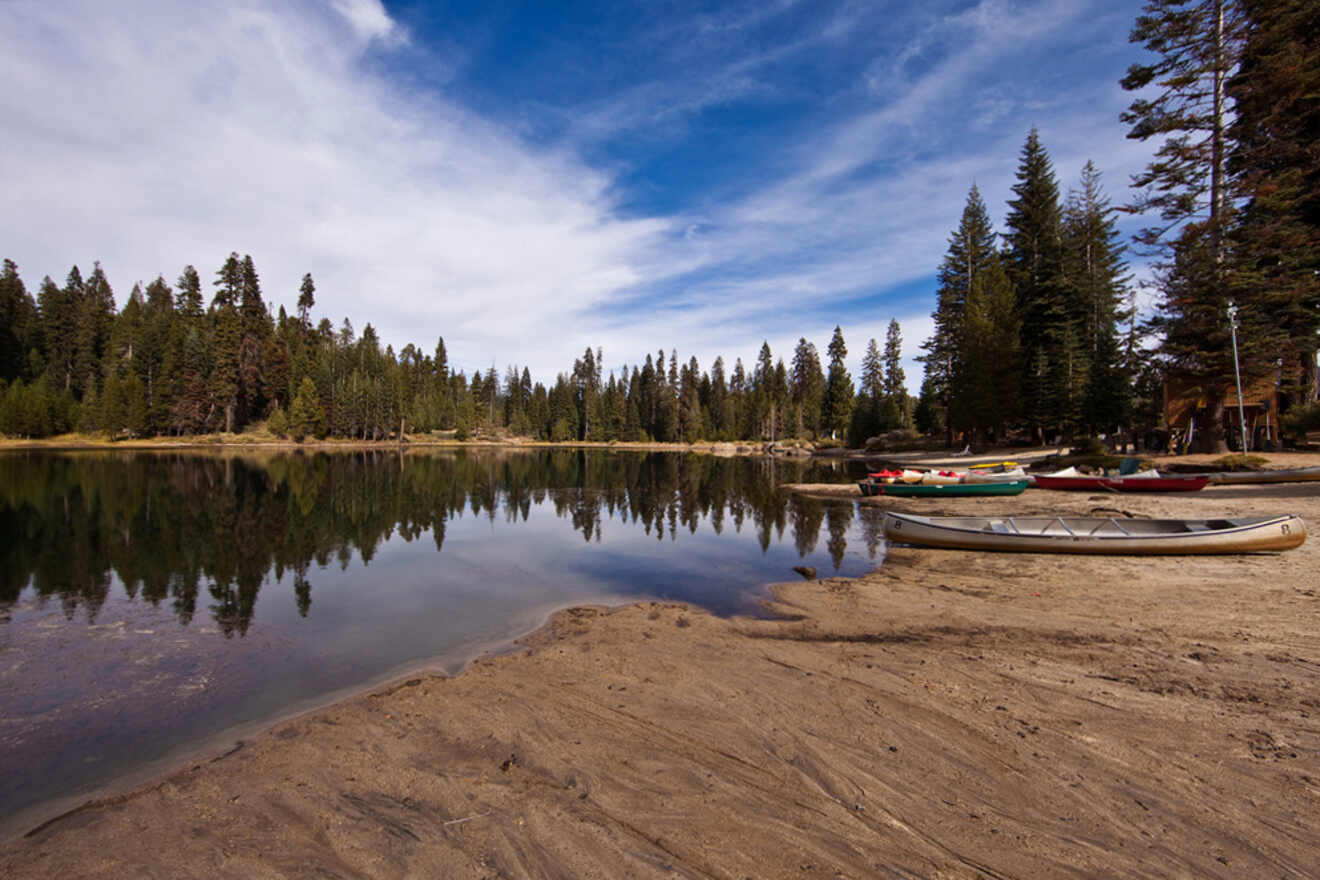 Several canoes on a sandy shore of a tranquil lake surrounded by trees under a partly cloudy sky.