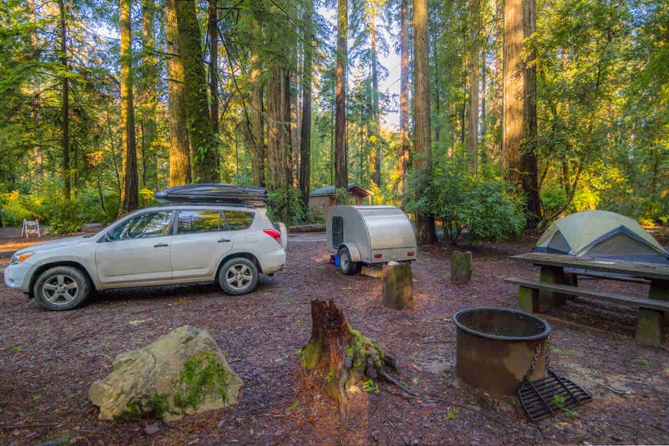 A campsite in a forest with a white SUV, a trailer, a tent, a picnic table, and a fire pit.