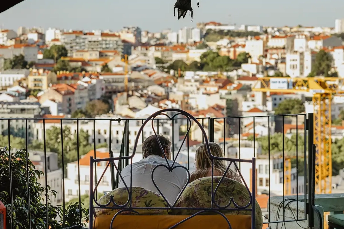 A couple sits on a balcony with a heart-shaped railing, overlooking an urban cityscape with numerous buildings and green spaces under a clear sky.