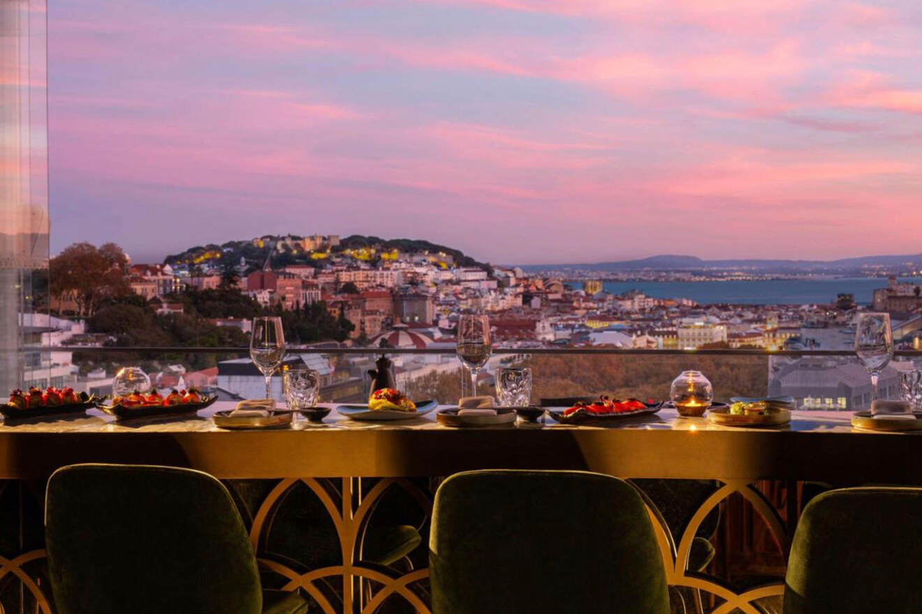 A dining table with plates and glasses overlooks a cityscape at sunset with a view of buildings, hills, and water in the background.