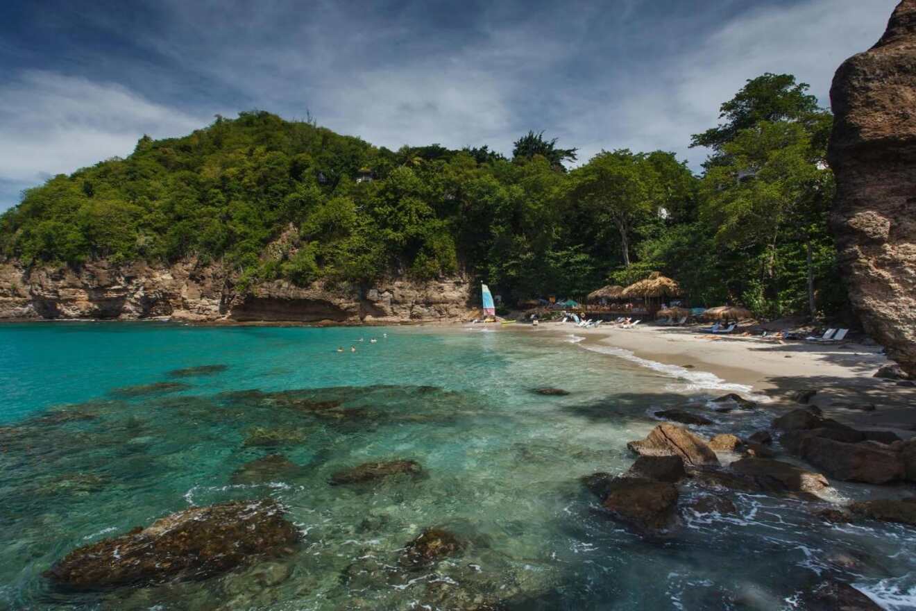 A tranquil beach scene with clear turquoise waters, a small secluded sandy beach, and lush green cliffs, under a partly cloudy sky.