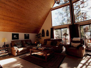 A cozy living room with wooden vaulted ceiling, large windows, and brown furniture including sofas and armchairs. Sunlight streams through the windows, illuminating the room.