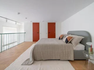 A bedroom with a double bed, beige bedding, pillows, and two red doors. There is a white rug on the wooden floor and a small round bedside table with a red book and lamp.