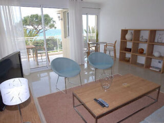 A modern living room with two blue chairs, a wooden coffee table, and a TV. The room features sliding glass doors leading to a balcony with a sea view and a dining area in the background.