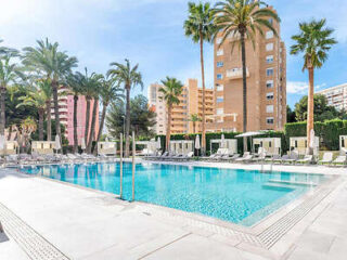 Outdoor swimming pool surrounded by lounge chairs, umbrellas, and tall palm trees with apartment buildings in the background.