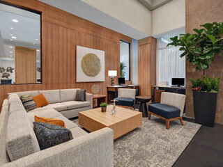 Modern lounge with a large beige sectional sofa, two blue chairs, wooden coffee table, tall plant, and wall art. A rug lies underneath, and a workspace with computers is visible in the background.