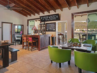 A cozy restaurant interior with wooden beams, green chairs, and tables. A chalkboard menu hangs above the bar counter, and large windows provide a view of the outside.