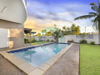 Outdoor swimming pool area next to a modern building with a shaded patio. Features a lawn, palm trees, and a colorful sky at sunset.