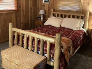 A cozy bedroom with wooden walls, a wooden bed frame, a red plaid blanket, two pillows, a wicker chest at the foot of the bed, and a bedside table with a lamp.