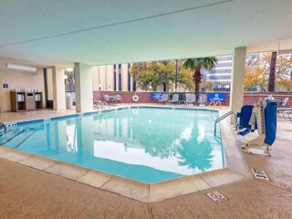 Indoor hotel swimming pool with accessible pool lift, lounge chairs, and a view of buildings and trees through large windows.