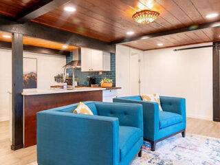 A modern living area with two blue armchairs on a patterned rug, a kitchen with teal backsplash tiles and a wooden ceiling with recessed lighting and a decorative ceiling light.