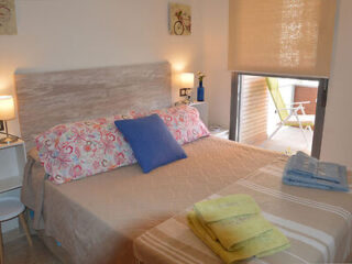 A neatly made bed with patterned pillows, a blue cushion, and folded towels at the foot. The room has soft lighting, wall art, and a window revealing an outdoor seating area with a lounge chair.