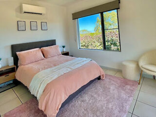 A bedroom featuring a neatly made bed with pink bedding, two side tables, a window with a view of greenery, a wall-mounted air conditioner, and a soft armchair.