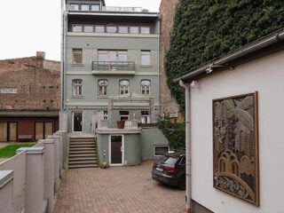 A multi-story gray building with multiple windows and a small balcony is seen from a courtyard containing a parked car and a wall-mounted art piece.