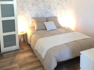 A neatly made double bed with grey bedding, a white throw blanket, and two illuminated bedside lamps against a patterned wallpaper. A sliding door is on the left, and a white dresser is partially visible.