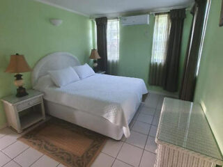 A simple and clean bedroom with pastel green walls, a white bed, wicker furniture, and windows letting in natural light.