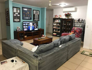 A living room with a U-shaped grey sofa facing a TV showing a man in a suit. The room has teal walls, framed pictures, a striped armchair, a bookshelf, and a coffee table.