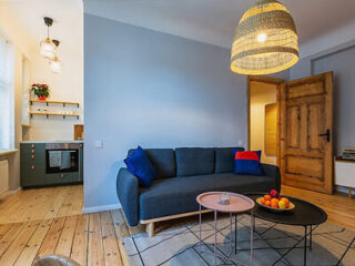 A modern living room with a dark gray sofa, blue and red cushions, a wooden coffee table, a large woven pendant lamp, and visible kitchen area in the background.