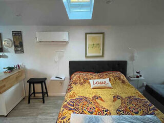 A bedroom with a bed covered by a colorful bedspread, a "Love" pillow, wall art, a skylight, a mini split AC unit, a small wooden counter, a stool, and wall-mounted lamps.