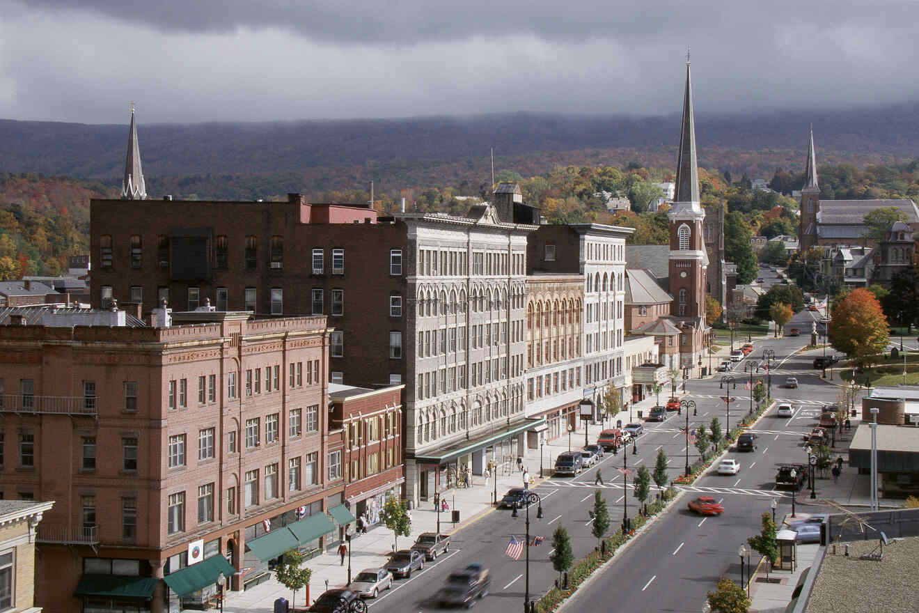 A small town street with historic buildings, shops, and churches with steeples, surrounded by trees and hills under a cloudy sky. Vehicles and pedestrians can be seen on the road and sidewalks.