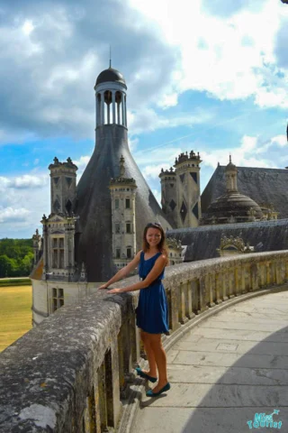 Young woman in a blue dress stands on a stone balcony overlooking the architectural details of a historic castle with round towers under a partly cloudy sky.