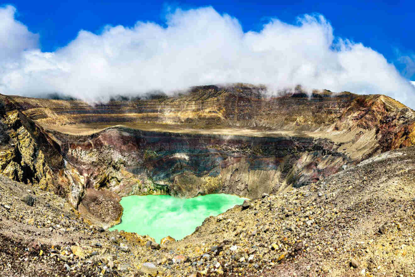 A volcanic crater with a green acidic lake at the bottom, rocky edges, and a partly cloudy sky overhead.