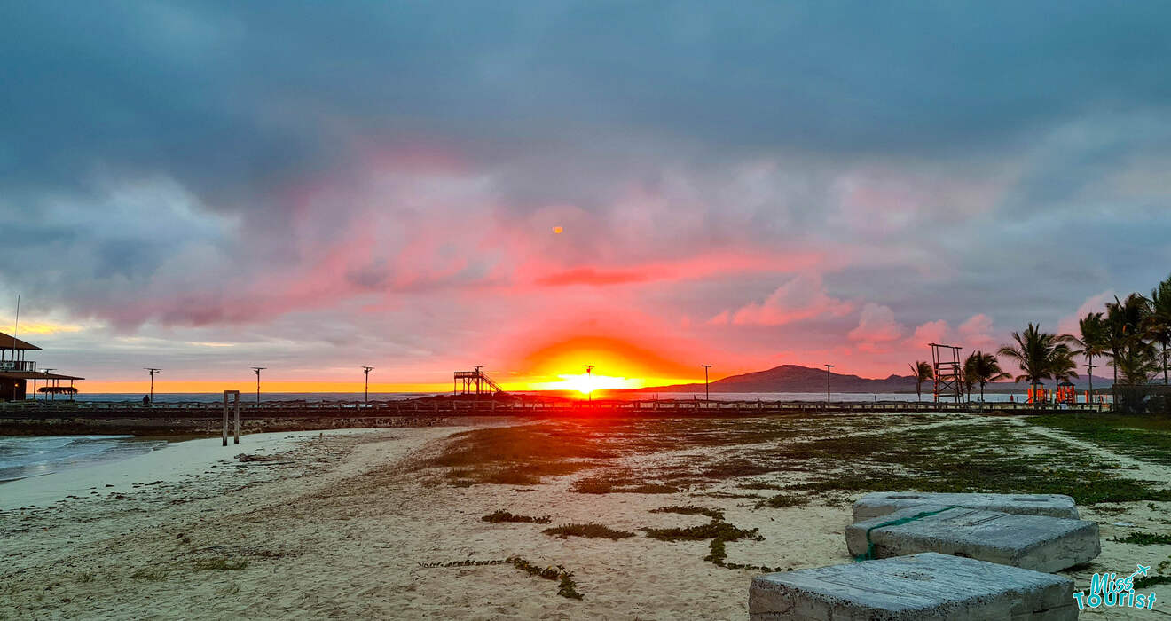 A serene beach scene at sunset with a partly cloudy sky. The sun sits just above the horizon, casting a vibrant glow over the sand, buildings, and palm trees in the background.