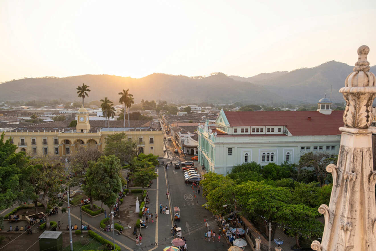 A cityscape view at sunset featuring historical buildings, a park with people, lined trees, and mountains in the background.