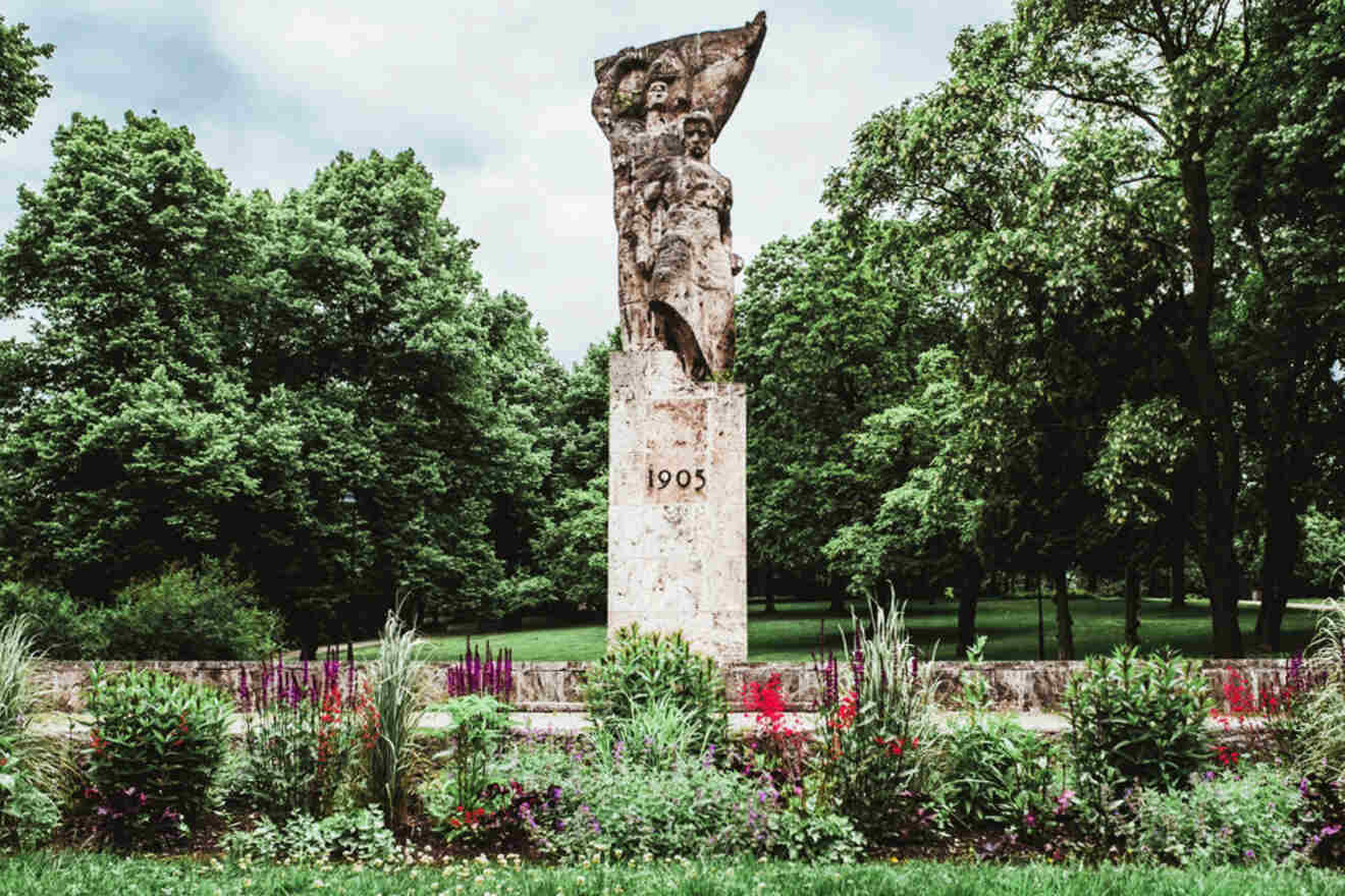 A stone monument inscribed with "1905" stands in a lush park, surrounded by various plants and flowers, with trees in the background.