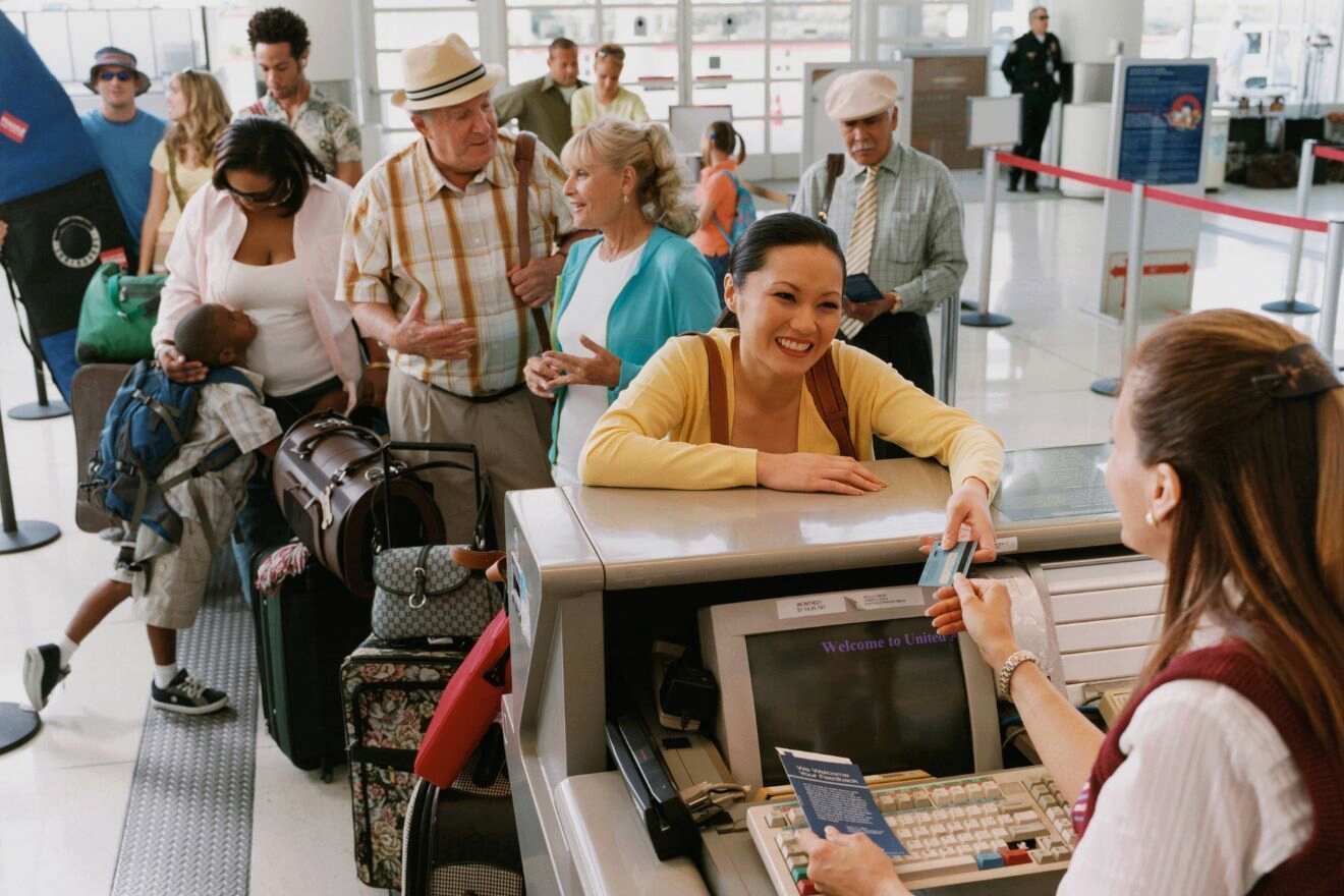 People are lined up at an airport check-in counter. A woman at the front of the line hands her ticket to an airline agent while others wait with their luggage.