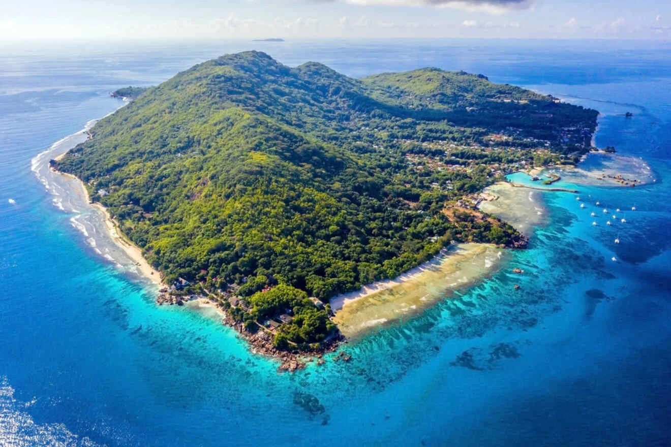 Aerial view of a lush green island surrounded by clear blue water, with scattered buildings along the coastline and several boats near the shore.