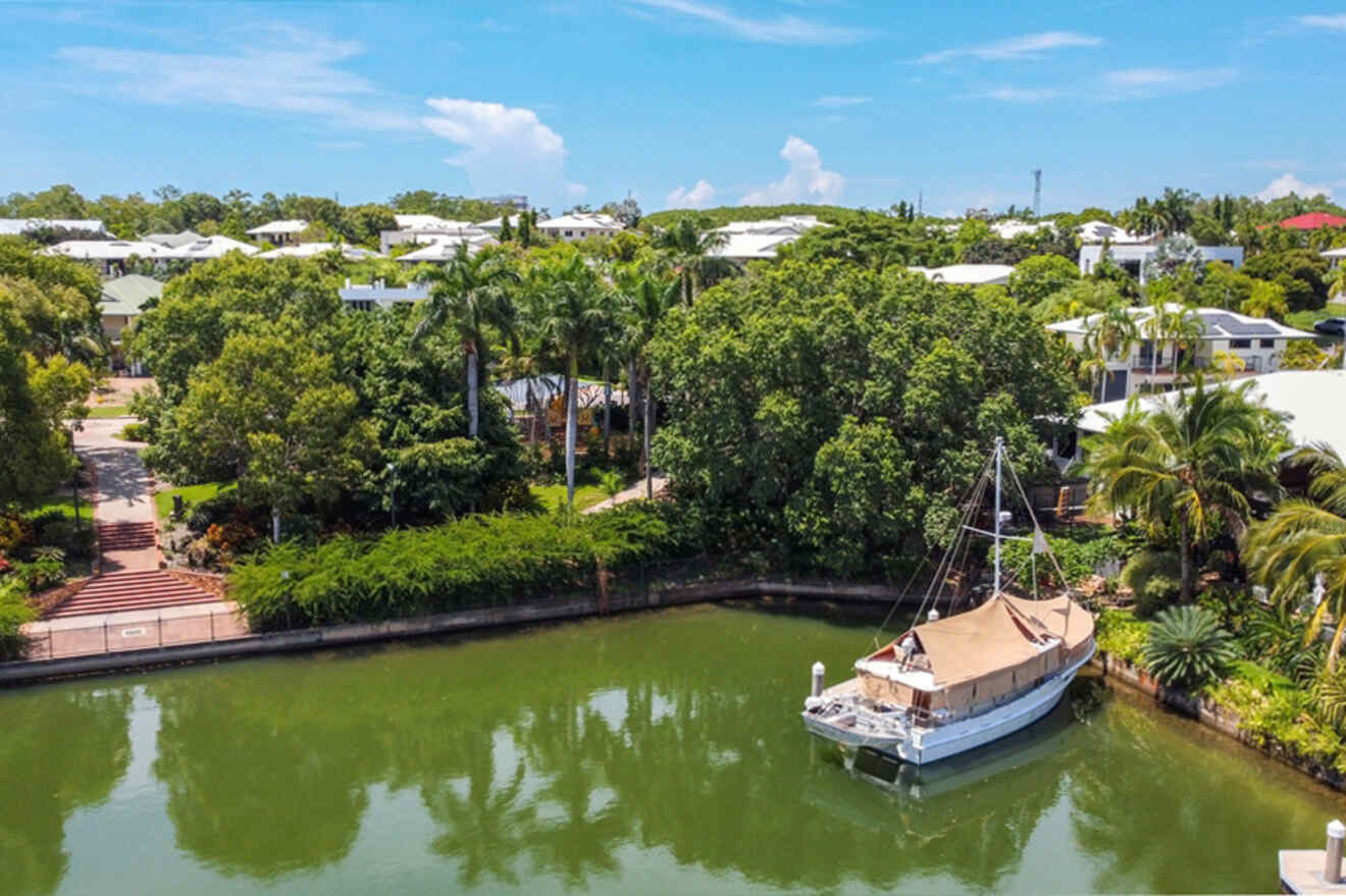 A boat docked on a calm canal with greenery, palm trees, and residential houses in the background under a blue sky.