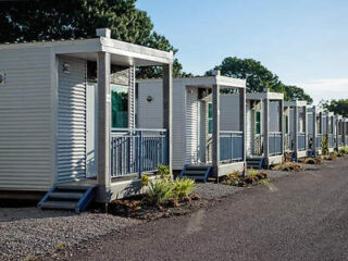 A row of identical tiny houses with small porches, set against a backdrop of trees and a clear sky. A gravel path runs alongside the houses.