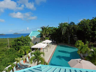 Aerial view of a pool area with umbrellas and lounge chairs surrounded by tropical greenery, overlooking the ocean and a clear blue sky in the background.