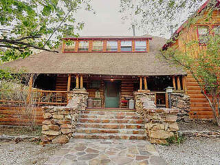 A rustic log cabin with a stone staircase leading to a covered porch. The cabin has two stories with windows and a wooden railing, surrounded by trees and vegetation.