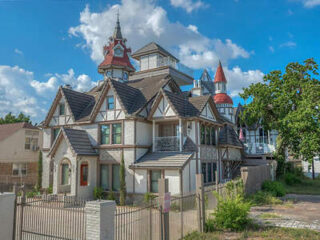 A large, multi-story house with intricate architectural details, including spires and gables, under a blue sky with scattered clouds. A metal fence surrounds the property.