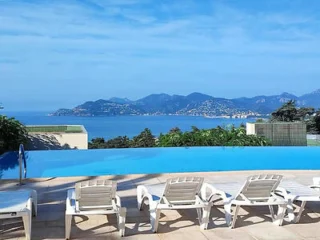 An infinity pool overlooking a scenic coastal view with mountains in the background. Five white loungers are arranged on the pool deck.
